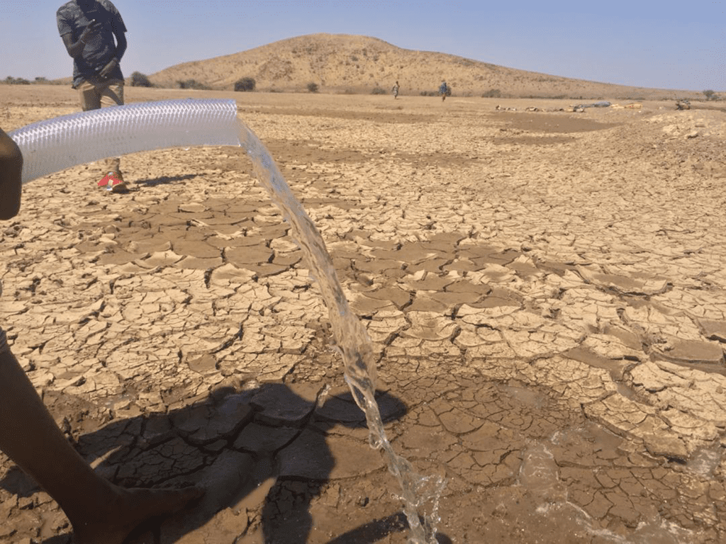 Water pouring out of a clear plastic pipe onto parched, draught-cracked land.