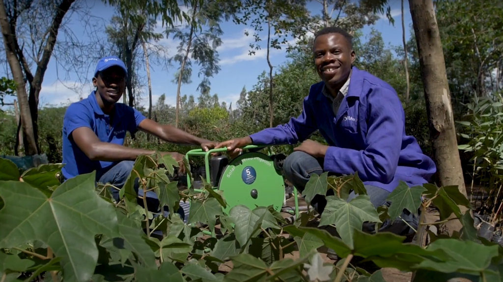 Two Futurepump technicians smile at the viewer as they sit with a Futurepump between them and green seedlings in the foreground