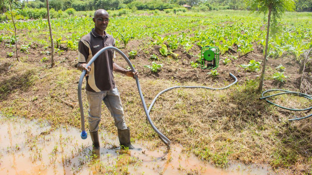 Farmer placing a hosepipe with a filter into his water source, which goes up to his ankles. In the background, we see a small SE1 and a hundred or so lettuce plants in the farm land. 