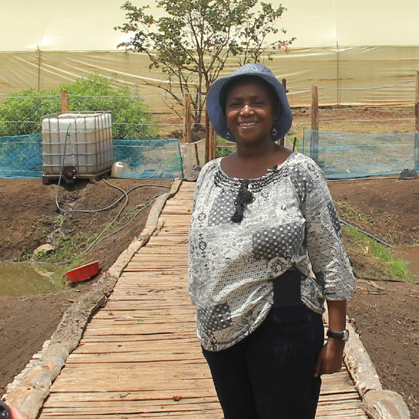 A smiling woman called Ann. She is standing on a wooden walkway on her farm. She has a hat on to protect from the bright sunlight and there is a white water storage tank behind her.