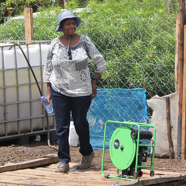 Ann is midstep, walking beside her SF2 solar water pump. Ann has a hat on in the bright sunshine, and a water bottle in her right hand.